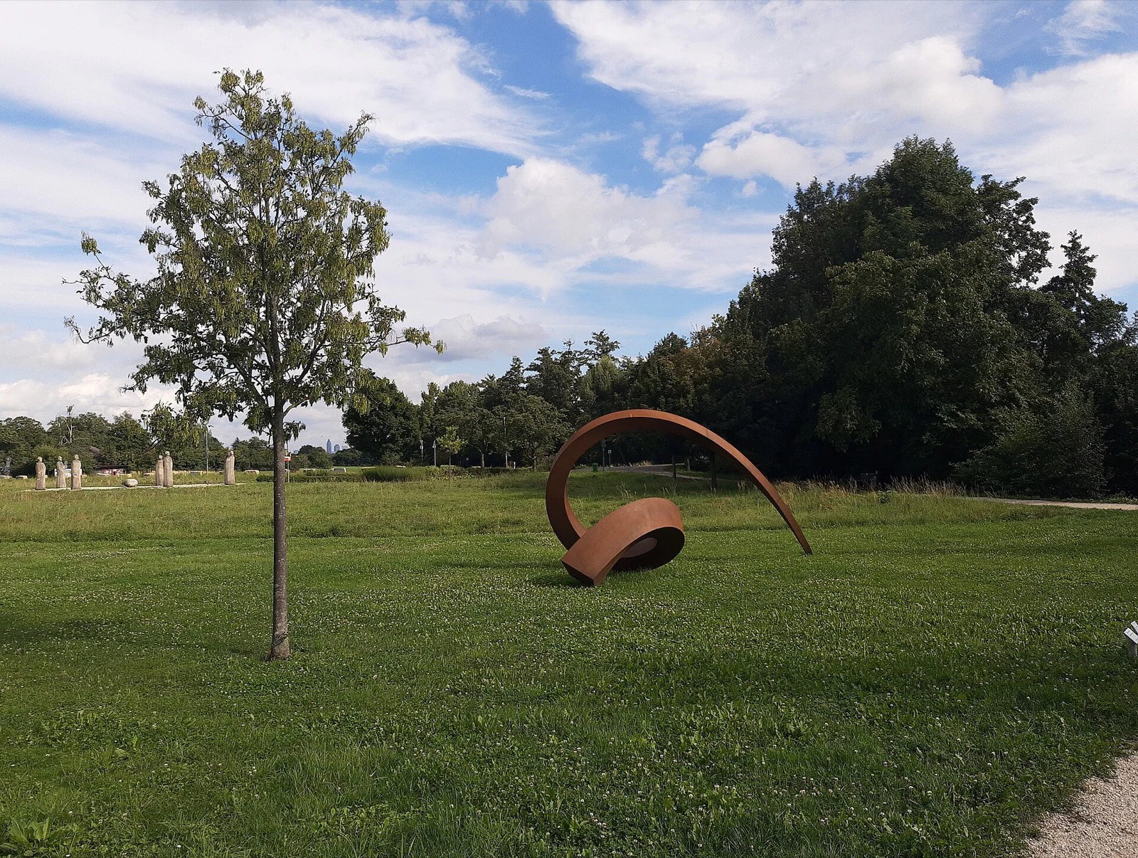 A curling weathering-steel sculpture on a park lawn, with a row of small stone figures visible in the distance.