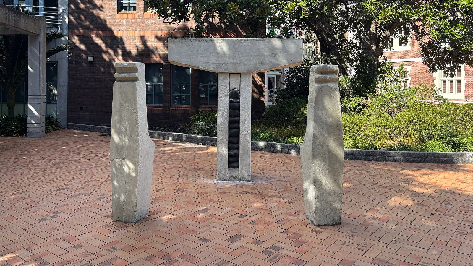 Three pale stone forms standing on a brick courtyard, with tree shadows falling in patches across their surfaces.