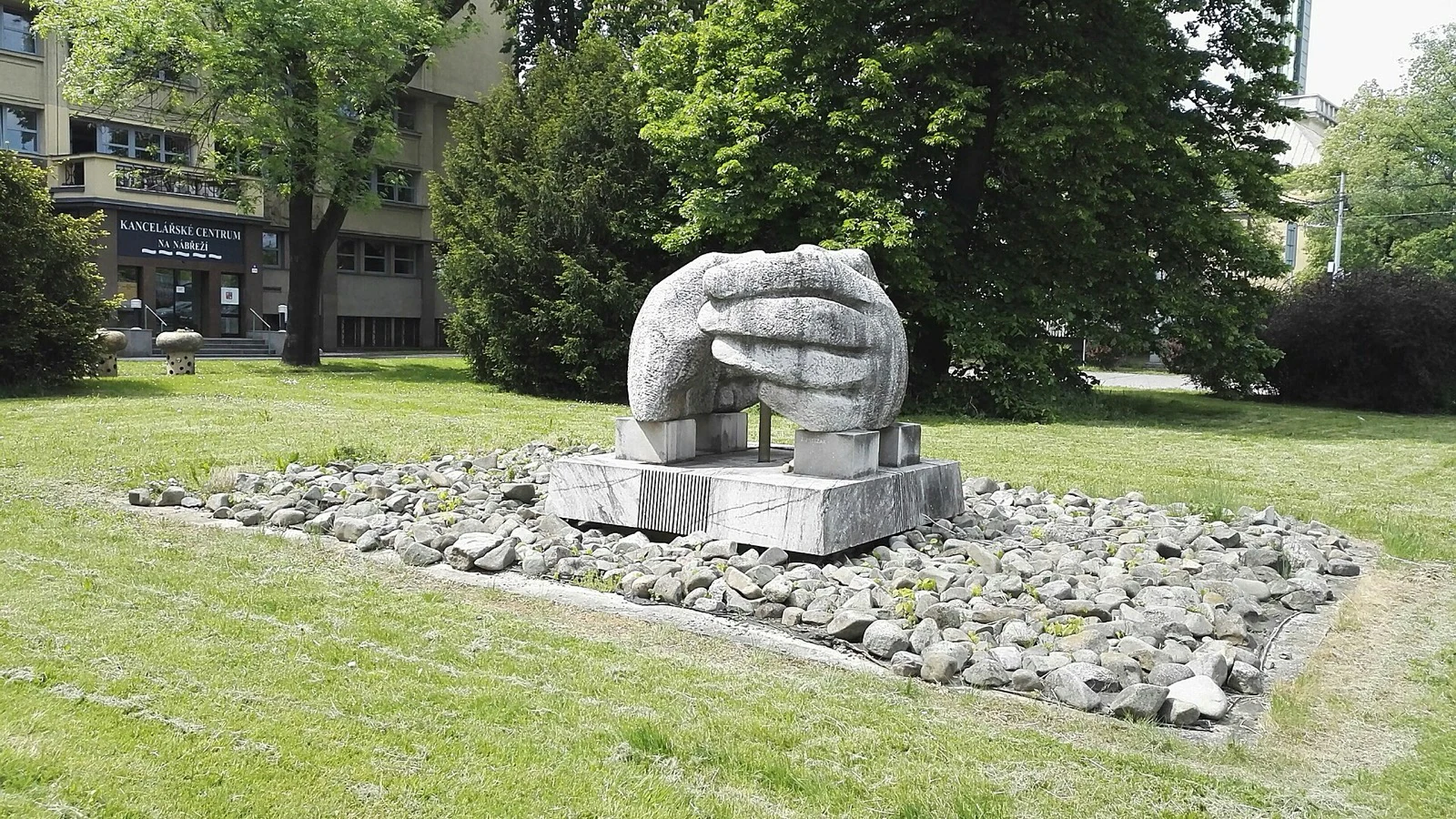A large stone sculpture of cupped hands on a plinth, set in a bed of river stones on a lawn beside an office building.