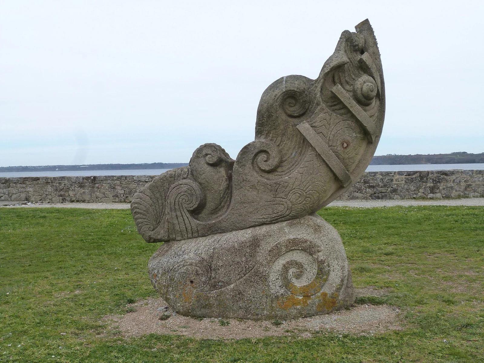 A large carved granite sculpture of curling wave forms on a coastal lawn, with orange lichen spreading across the base.