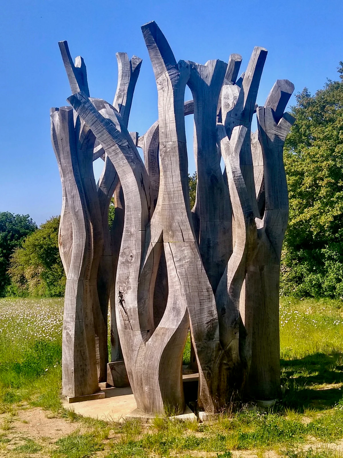 Tall carved oak columns with inscribed text standing together in a wildflower meadow under a clear sky.