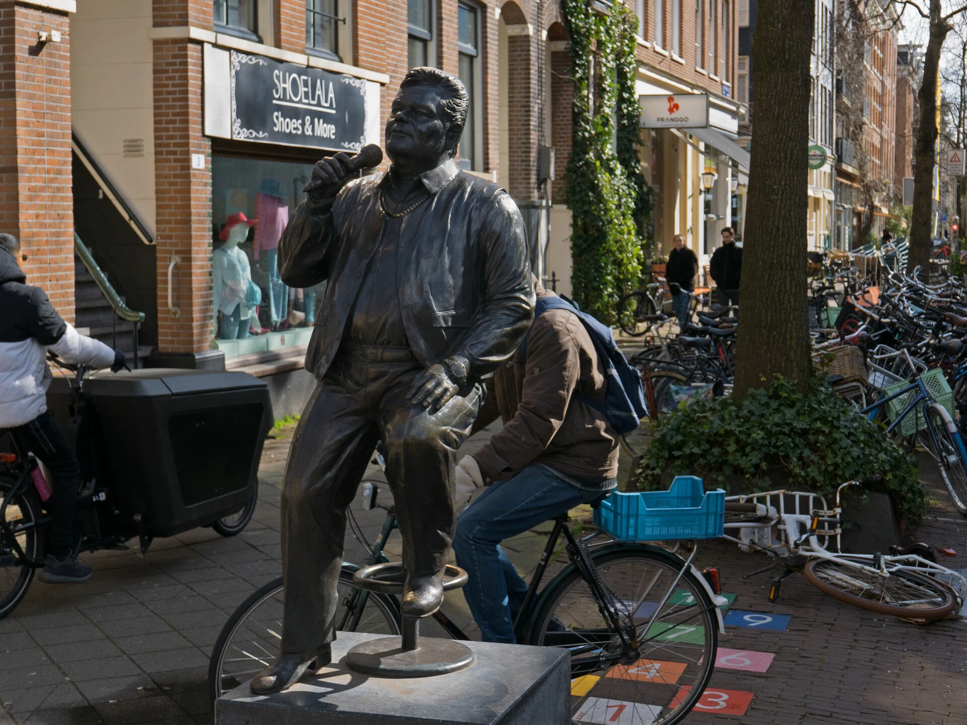 Cyclists passing a bronze sculpture of a seated man on a narrow Amsterdam sidewalk in morning light.