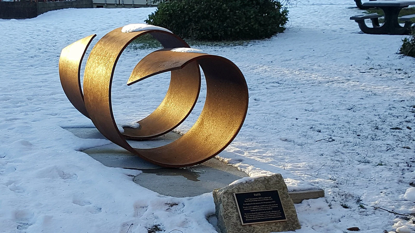 A curling weathered-steel sculpture lit gold by low sun on fresh snow, with a small dedication plaque on a stone block nearby.