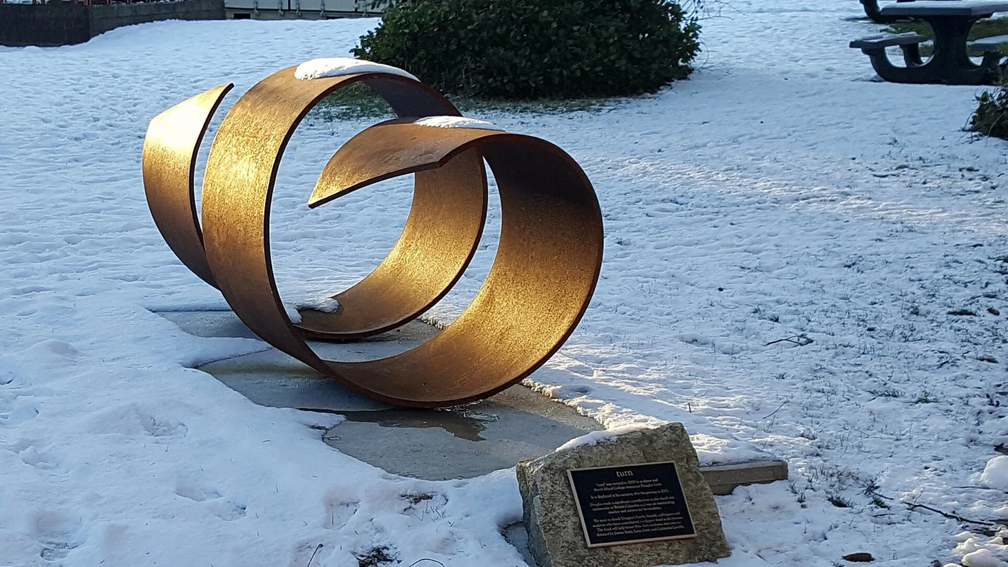 A curling weathered-steel sculpture lit gold by low sun on fresh snow, with a small dedication plaque on a stone block nearby.