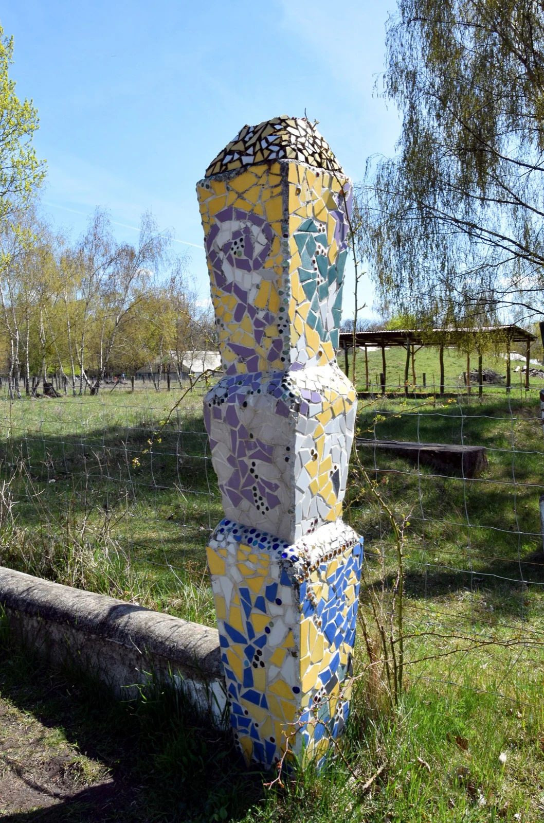 A tall column covered in yellow, blue, purple, and white mosaic tile fragments standing in a grassy park.