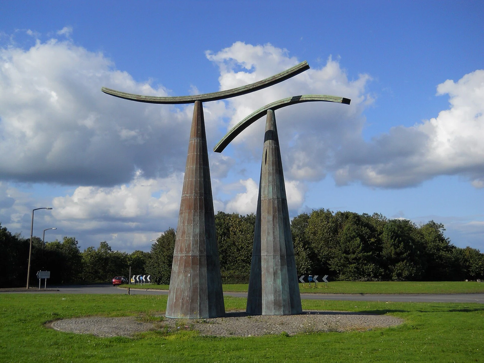 Two tall bronze columns supporting curved arms, installed on a grass roundabout encircled by road.