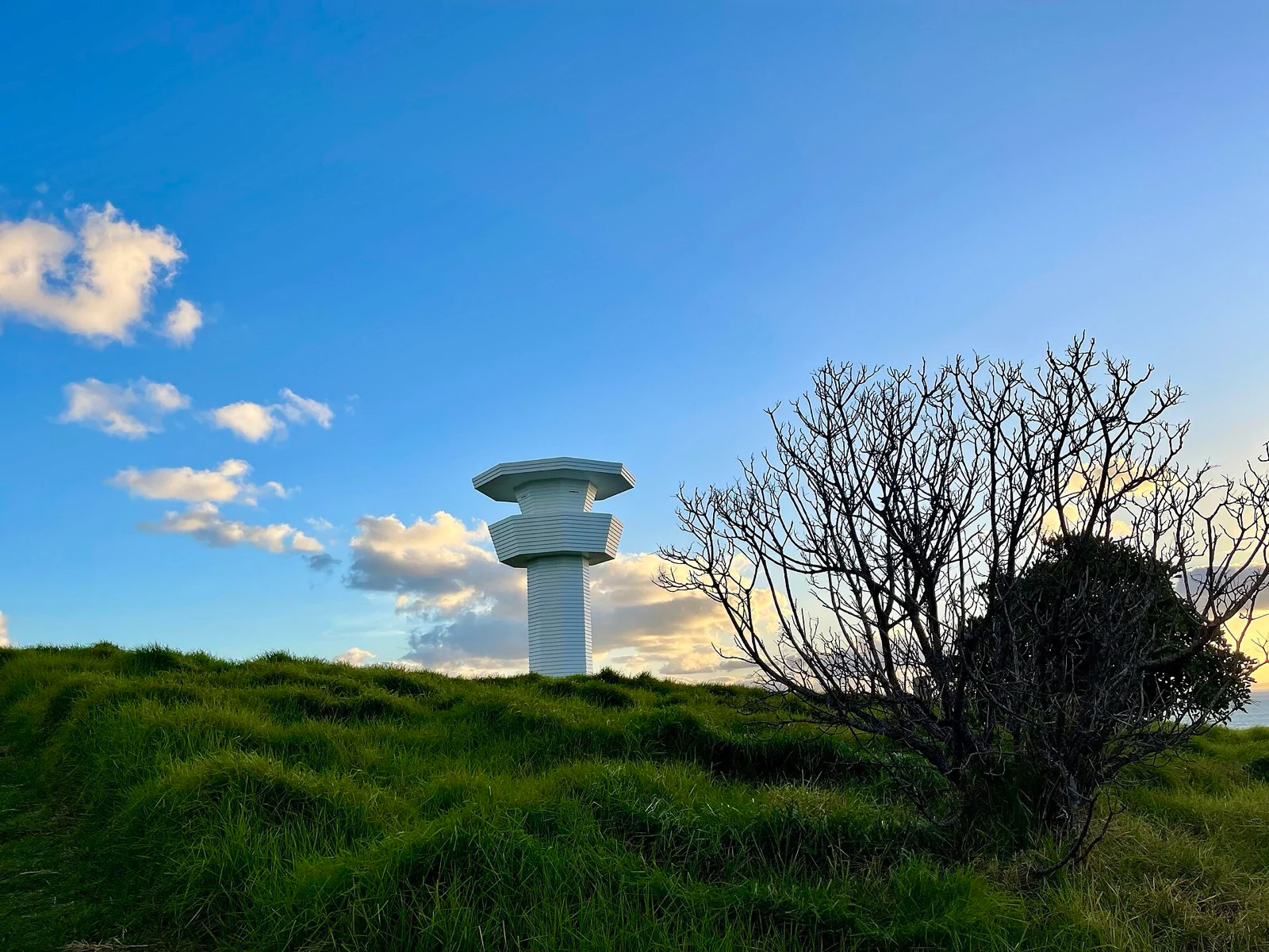 A tall white sculpture resembling a stacked tower standing on a grassy headland against an evening sky.