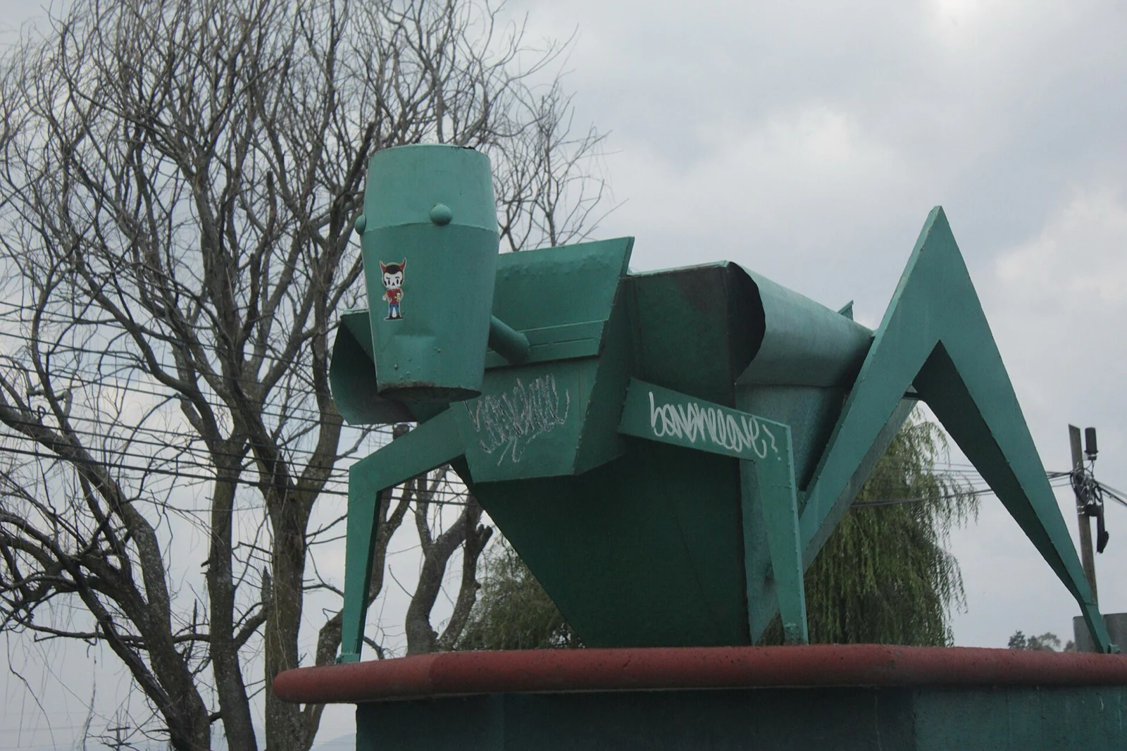 A large green painted metal grasshopper sculpture on a plinth against an overcast sky.