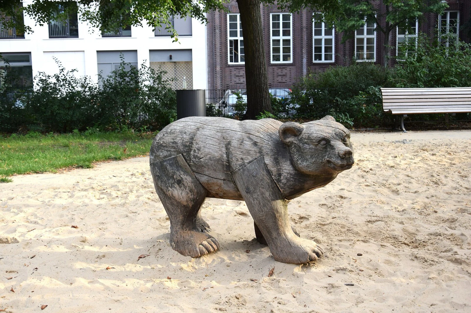 A carved wooden bear standing in a sandpit in a city park, with benches and buildings behind.