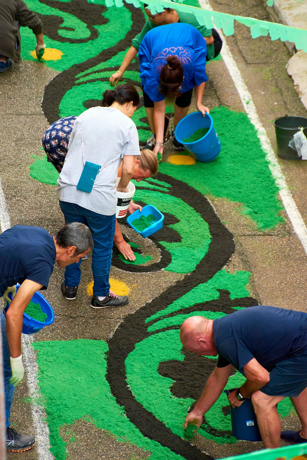 Volunteers kneeling in a closed street, filling a large scrolling pattern with green sawdust from buckets