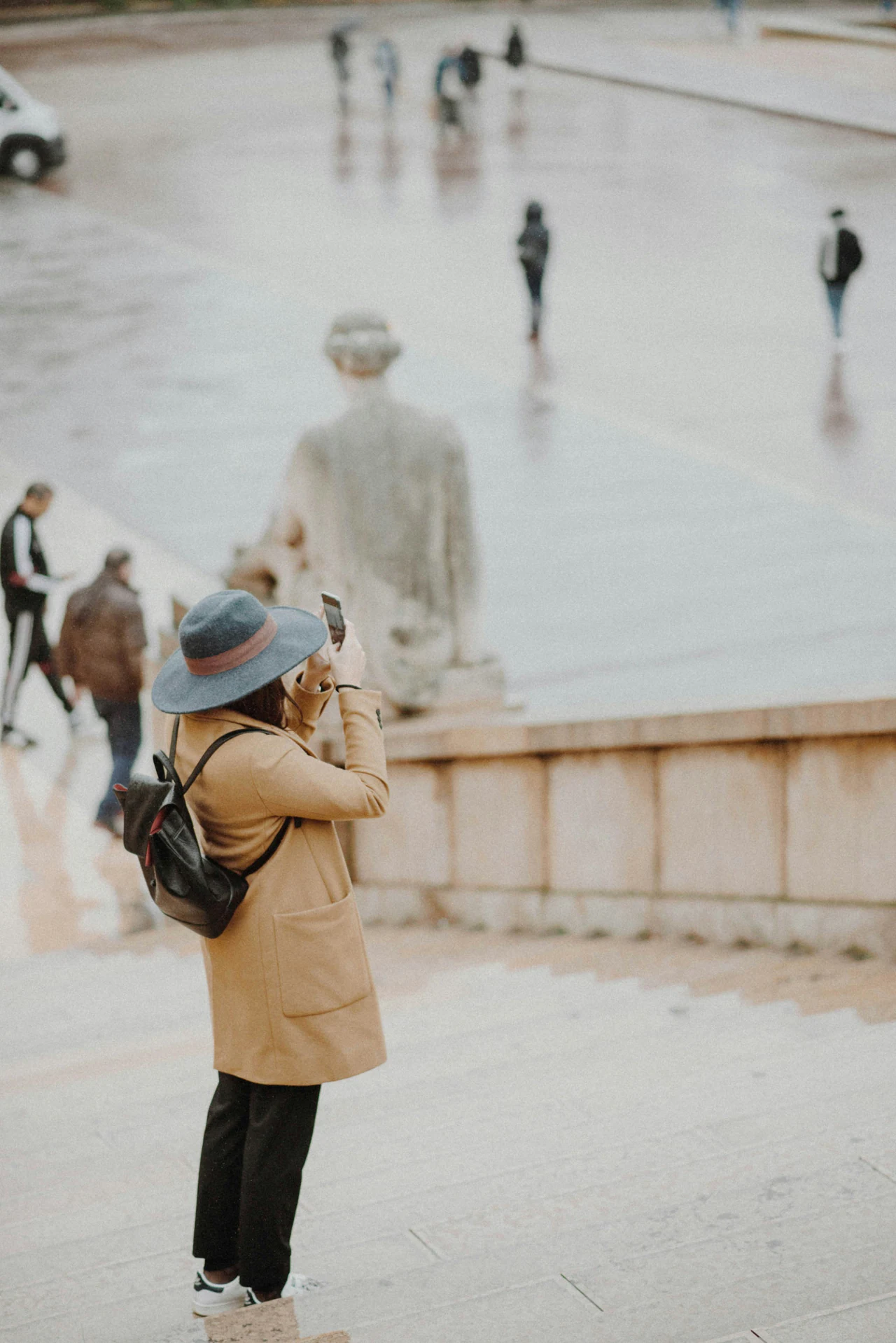 A visitor in a wide-brimmed hat photographing a stone monument across a rain-wet public square