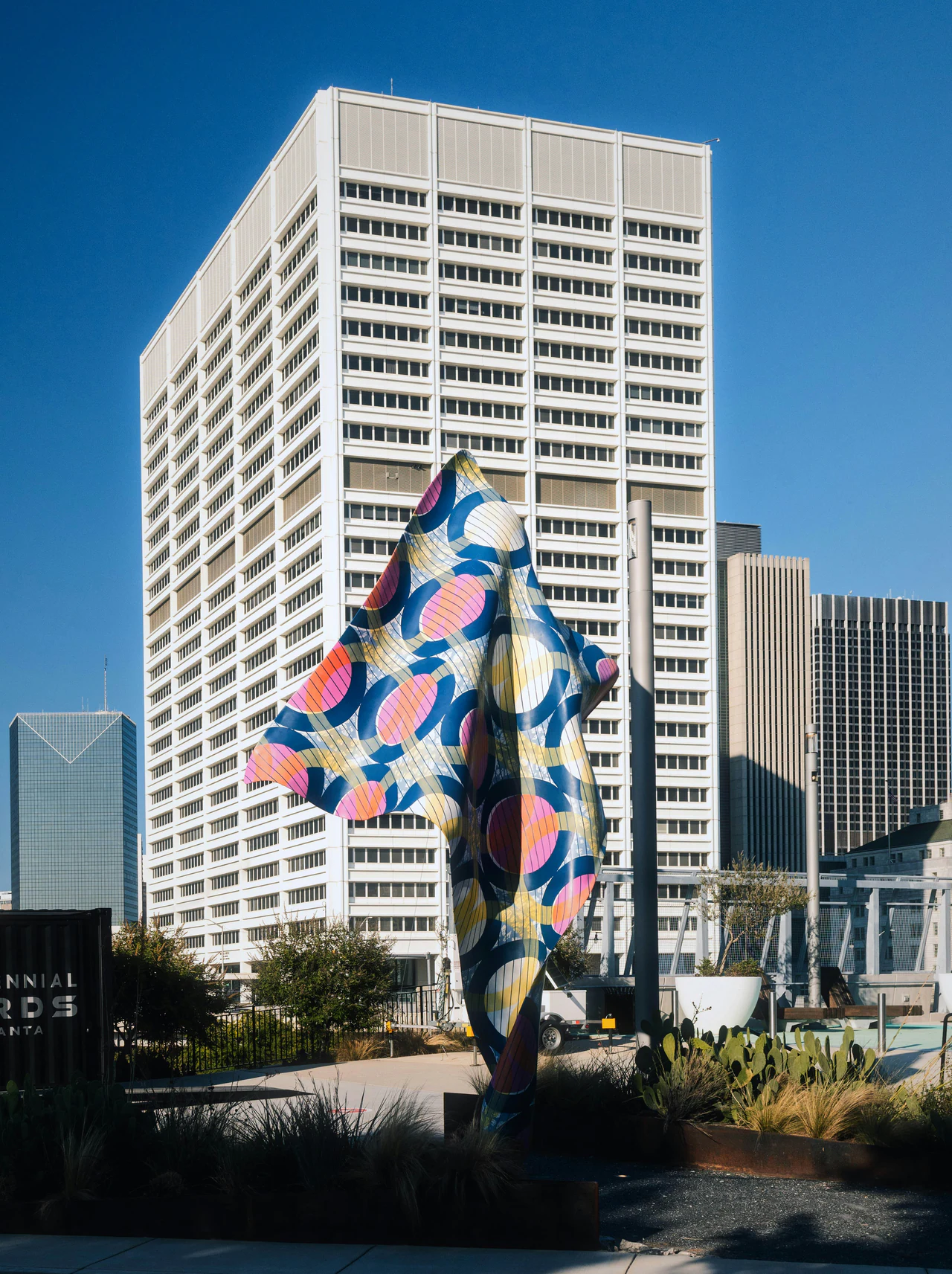 A tall sculpture patterned in pink, orange and blue circles standing in a downtown plaza below office towers
